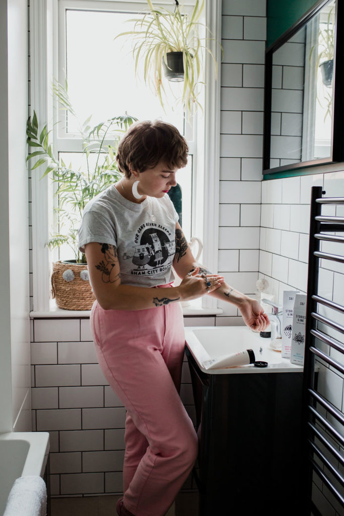 Grace is applying cream to her tattooed arm in her friend's gorgeous bathroom. white tiles and pink trousers are a winning combo.