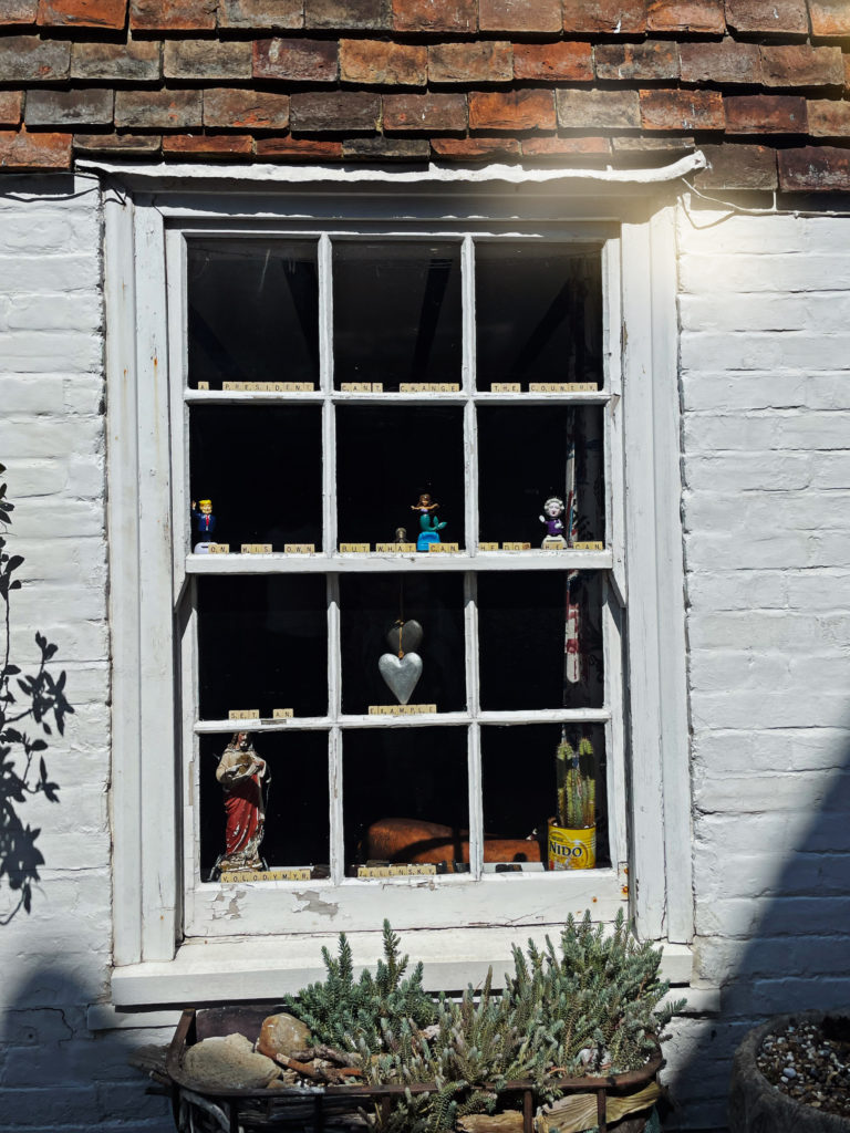 A very beautiful white window set into white painted brick, on an old fashioned street in Rye. There are ornaments, hanging decorations and Scrabble tiles spelling out some kind of message in the window.