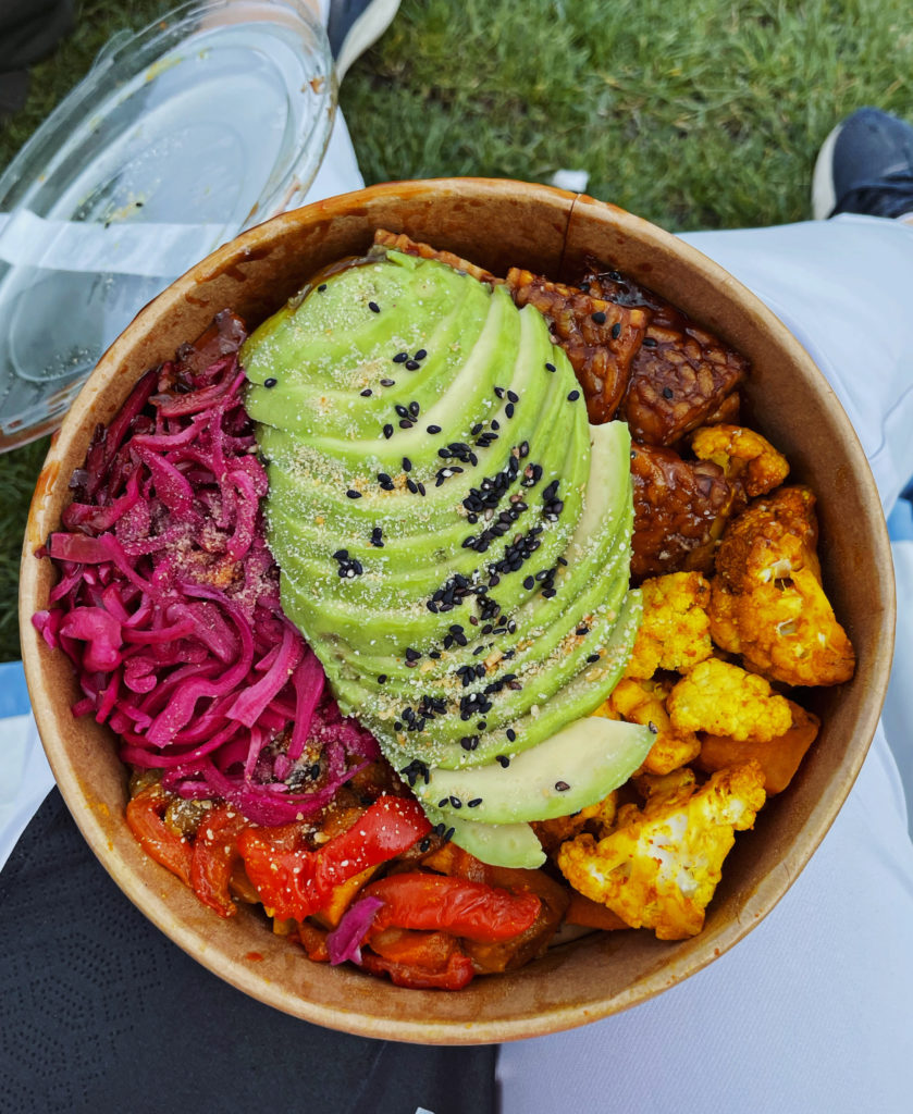 a takeaway paper bowl of avocado, tempeh and vegetables.