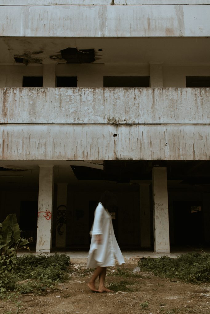 a woman in a long white shirt, blurred, walking past an abandoned building.